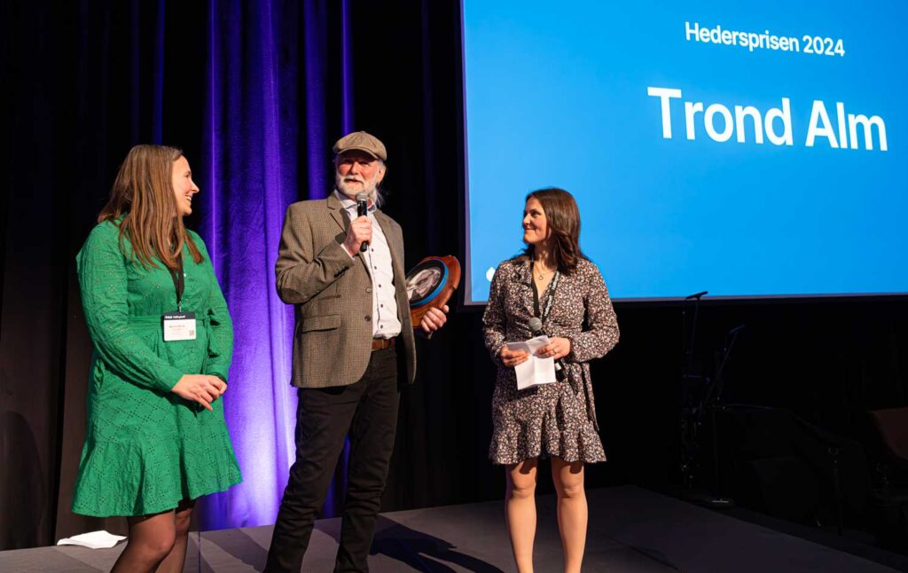 Trond Alm fikk Hallingtreffs hederspris 2024. Her med møtelederne Martina Bergh Svedahl (t.v.) og Borghild T. Folkedal.